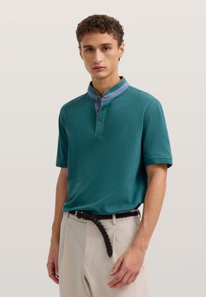 Junger Mann trägt ein türkisfarbenes kurzärmeliges Poloshirt mit kontrastierendem Kragen und beige Hosen mit einem dunklen geflochtenen Gürtel, steht vor einem einfarbigen Hintergrund.
