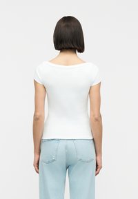 Γυναίκα με ίσια καστανά μαλλιά που φοράει εφαρμοστή λευκή κοντομάνικη μπλούζα και ανοιχτό μπλε ψηλόμεσο τζιν, με την πλάτη στραμμένη σε λευκό φόντο.