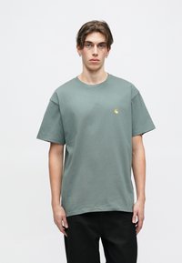 Grøn bomulds t-shirt med rund halsudskæring, korte ærmer og et lille guld-logo på brystet. Stoffet ser blødt og letvægts ud.