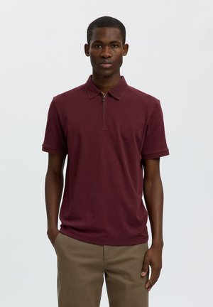 Giovane uomo che indossa una polo bordeaux a maniche corte con cerniera frontale e pantaloni beige, in piedi davanti a uno sfondo chiaro e uniforme.