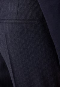Tmavě modré přiléhavé kalhoty s jemným vertikálním pruhovaným vzorem. Obsahují zadní kapsu s elegantním otvorem. Materiál se jeví jako hladký a odolný.