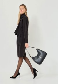 Chaqueta negra corta sobre una falda negra, combinada con medias negras transparentes y tacones altos negros. Sostiene un bolso estructurado negro con acentos dorados.