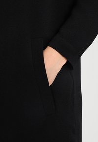 Svart tygplagg med en närbild av en hand som sträcker sig in i en sidoficka; slät textur och enkel design utan synliga mönster.