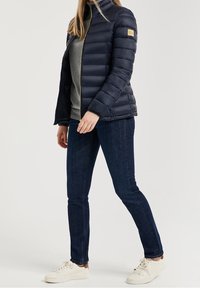 Marinblå puffjacka med hög krage, gulddragkedja och patch-logotyp; kombinerad med en grå topp, mörka jeans och vita sneakers.