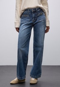 Jeans in denim blu a gamba larga con una finitura sbiadita, dotati di hardware in tonalità oro, due tasche frontali e una tradizionale chiusura a bottoni.