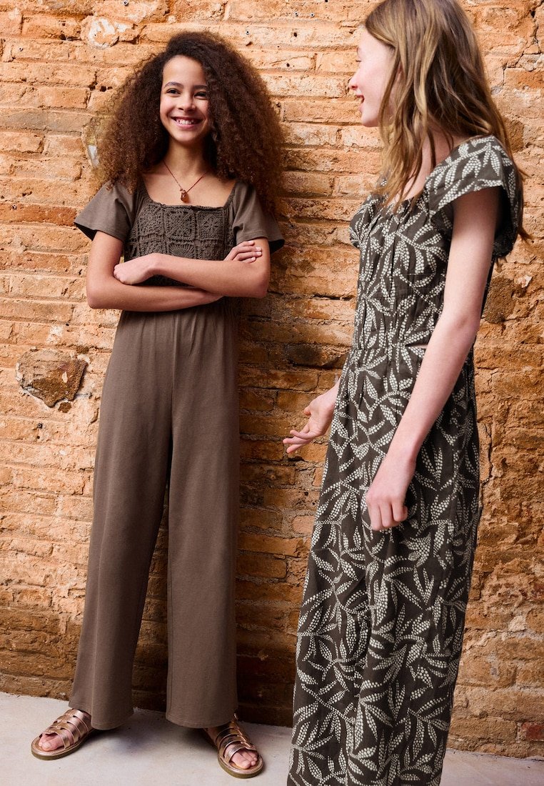 Deux filles se tiennent debout et discutent devant un mur en briques texturé, l'une en combinaison marron avec des cheveux bouclés, l'autre en robe à motifs avec des cheveux lisses.