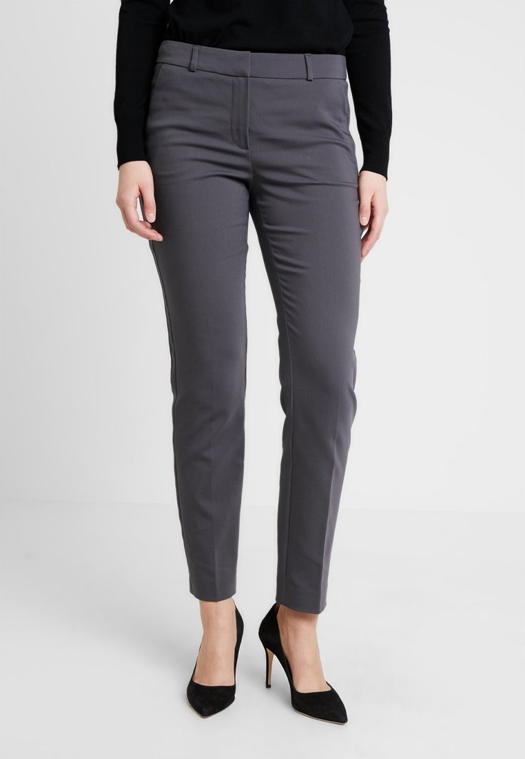 Pantalon gris sur mesure à coupe slim, avec un devant plat, des passants pour ceinture et des poches latérales. Associé à des chaussures noires à talons hauts.