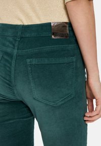 Dark green corduroy pants with a smooth texture. Features two back pockets and a metallic brand tag on the waistband. Close-up angle.