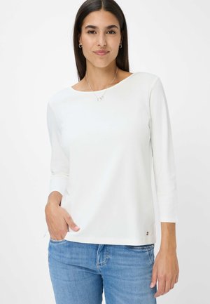 Top bianco a maniche lunghe con scollo a barca, texture morbida e un dettaglio logo vicino all'orlo, abbinato a jeans di denim blu.