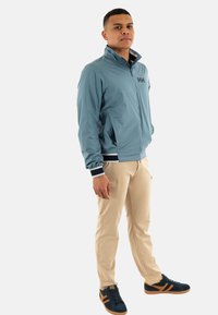 Giacca azzurra chiara con zip e polsini elasticizzati, abbinata a pantaloni beige e sneakers scure con accenti arancioni; il modello è in posizione frontale.