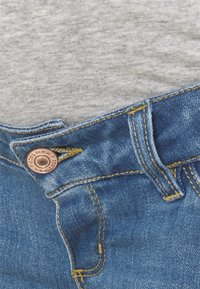 Jeans de mezclilla azul con un botón de cobre, costuras amarillas y una camiseta de algodón gris visible por encima de la cinturilla.