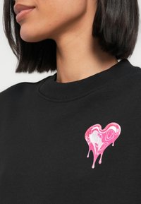 Μαύρο φούτερ με πολύχρωμο σχέδιο καρδιάς που στάζει σε αποχρώσεις του ροζ, με χαμογελαστά πρόσωπα και ριμπ λαιμόκοψη.