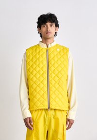 Veste matelassée jaune avec fermeture éclair sur le devant, comportant un col montant et un petit logo sur la poitrine. Portée par-dessus une chemise crème.