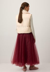 Bēšs pūkains veste ar augstu apkakli, uzvilkta pār garu bordo tilla svārku. Modelis valkā rozā apavus, mati sasukti atpakaļ. Vienkāršas, gludas faktūras.