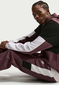 Jeune personne aux cheveux tressés assise en tailleur, portant un survêtement Nike bordeaux, noir et blanc avec capuche, regardant vers la caméra.