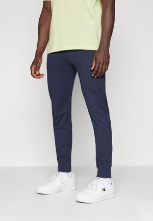 Mann trägt marineblaue Jogginghosen, weiße Champion-Sneaker, weiße Socken und ein hellgelbes T-Shirt und steht auf einem schlichten hellgrauen Boden.