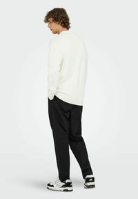 Junger Mann mit lockigem Haar steht nach links schauend, trägt einen weißen Pullover, schwarze Hosen und schwarz-weiße Sneaker vor einem schlichten Hintergrund.