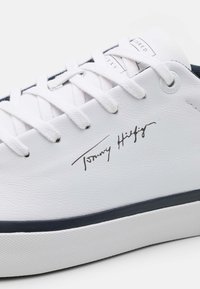 Bílé kožené tenisky s námořnickými akcenty, vybavené plochými bílými tkaničkami a s podpisem "Tommy Hilfiger" tištěným hnědou barvou na boku.