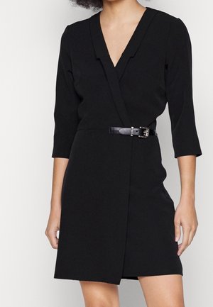 Frau trägt ein schwarzes Wickelkleid mit dreiviertellangen Ärmeln und einem dünnen schwarzen Gürtel mit dekorativer Schnalle in der Taille.