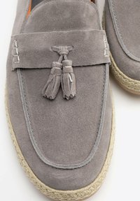 Mocasines de ante gris con flecos decorativos, que presentan costuras claras y un borde de yute trenzado alrededor de la suela. Textura suave y diseño casual.