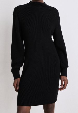 Czarna sukienka dzianinowa z prążkowanej dzianiny z długimi bufkowymi rękawami, noszona przez stojącą osobę, widoczna od ramion do połowy uda na jednolitym tle.