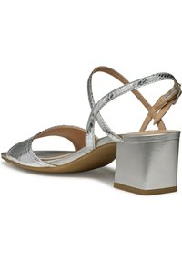 Sandalias plateadas de tacón bajo que presentan un upper texturizado y brillante con diseño de punta descubierta, correas en el tobillo entrelazadas y un tacón cuadrado bajo.