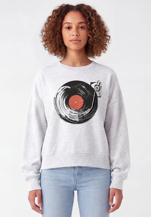 Jeune femme aux cheveux bouclés portant un sweat-shirt gris clair avec un motif de disque vinyle noir et rouge et platine, associé à un jean bleu clair.