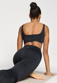 Μαύρο αθλητικό σουτιέν και κολάν, εφαρμοστός σχεδιασμός, λεία υφή, με ελάχιστες ραφές, με λογότυπο στη ζώνη μέσης. Το μοντέλο σε θέση τεντώματος.