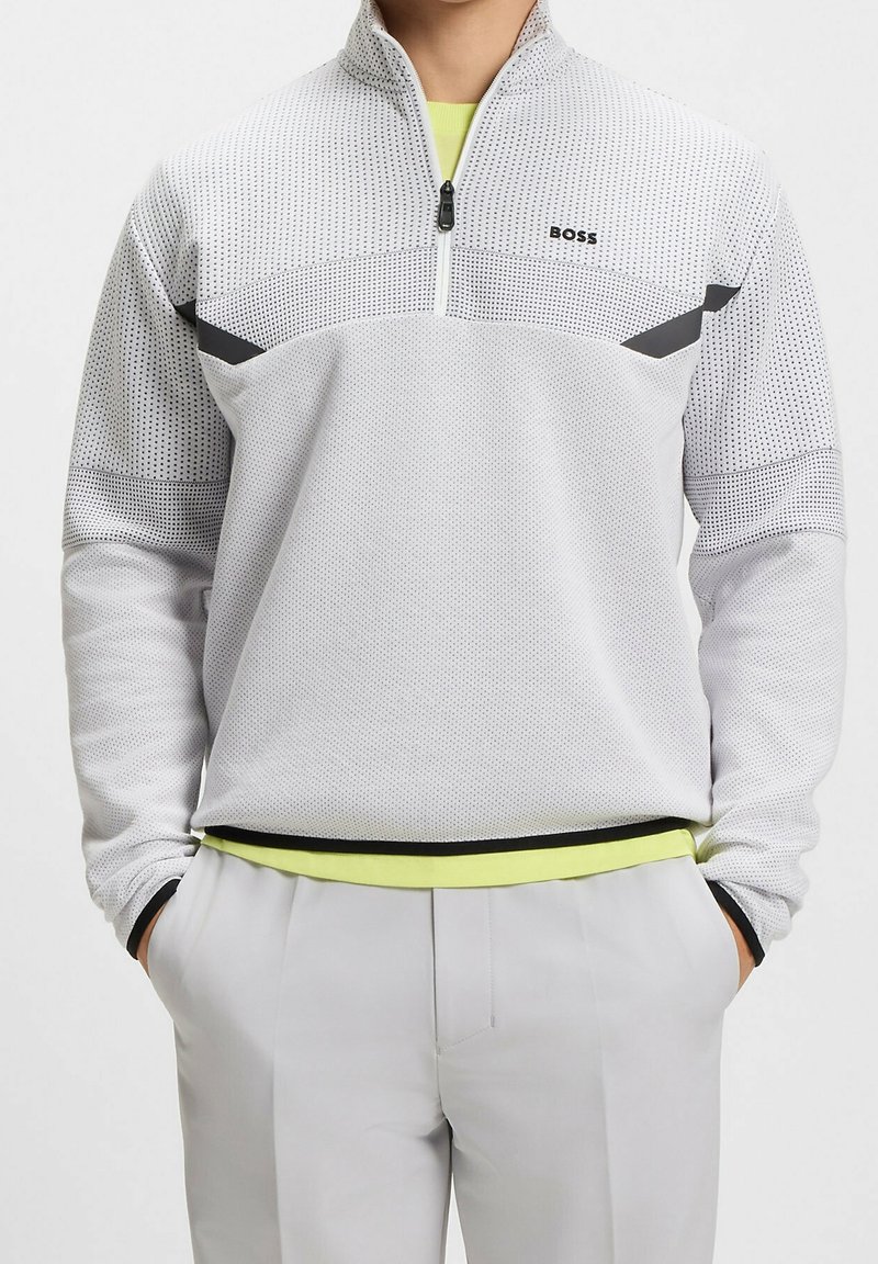 Personne portant un pull gris clair avec des accents noirs et le logo "BOSS", associé à un pantalon gris clair et les mains dans les poches.
