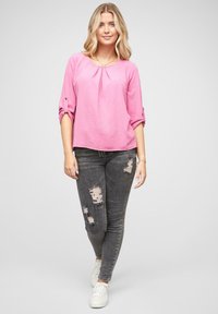 Blusa rosa con texture, scollatura tonda e dettagli plissettati, abbinata a jeans skinny grigi scuri con accenti strappati e sneakers bianche.