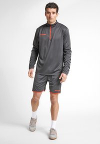 Grå atletisk långärmad topp med kort dragkedja och orange accenter, i kombination med grå shorts med orange kant, bärs med ljusgrå sneakers.