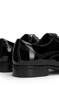 Zapatos de vestir de charol negro con detalles de brogue, superficie lisa, tacón bajo y cordones. Presentan una punta redondeada y acentos cosidos.