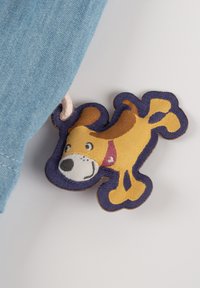 Blaues Gewebe mit einem farbenfrohen, gestickten Hundemarke in Gelb und Braun mit lila Umrandung, die an einem Kleidungsstück befestigt ist.