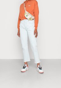 Casual outfit with an orange hoodie, light blue high-waisted jeans with frayed hem, and multicolored sneakers. Fanny pack features geometric patterns.