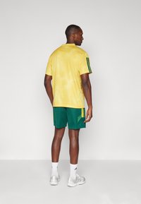 Rumena športna majica z teksturiranim vzorcem, zelena kratka hlača in tri zelene črte na ramenih, oblečena z belimi nogavicami in sivimi športnimi čevlji.