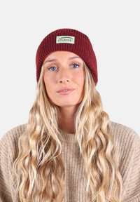 Bonnet en maille côtelée bourgogne avec une étiquette rectangulaire verte portant le texte "OXBOW". Le matériau semble doux et s'adapte confortablement.