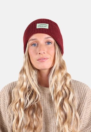Bonnet en maille côtelée bourgogne avec une étiquette rectangulaire verte portant le texte "OXBOW". Le matériau semble doux et s'adapte confortablement.