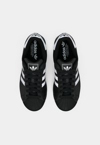 Black sneakers with white three-stripe design, rubber toe cap, and lace-up closure. Features branded tongue and heel accents.