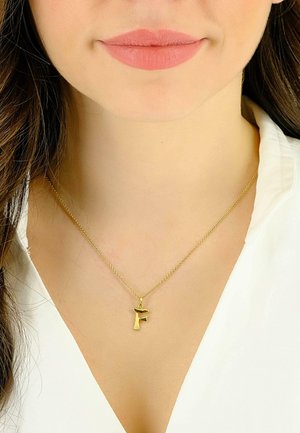 Collana con catenina d'oro e pendente decorativo a forma della lettera "F". Il pendente ha una finitura liscia e riflettente. Indossato su una maglietta bianca.