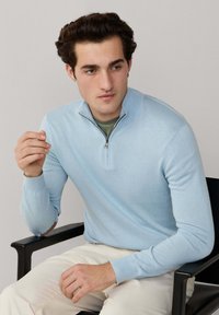 Jeune homme aux cheveux bouclés foncés assis sur une chaise noire, portant un pull à demi-fermeture éclair bleu clair et un pantalon crème, regardant légèrement sur le côté.