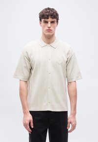 Νέος άνδρας με σγουρά μαλλιά, φορώντας μια μπεζ κοντομάνικη πουκάμισο με γιακά και μικρό έμβλημα στο αριστερό στήθος, συνδυασμένο με μαύρο παντελόνι.