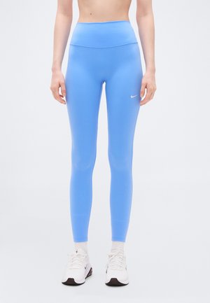 Femme portant un legging de sport bleu taille haute et des baskets blanches Nike, debout devant un fond blanc uni.