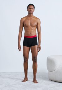 Μαύρα ανδρικά εσώρουχα boxer με κόκκινη ζώνη μέσης "HUGO", με στενή εφαρμογή και απαλό βαμβακερό υλικό. Το μοντέλο στέκεται ξυπόλητο μπροστά σε ανοιχτό φόντο.