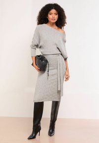Hellgraues Strickkleid mit schulterfreiem Design, strukturiertem Muster, Stoffgürtel und einer schwarzen Clutch. Kombiniert mit hohen, schwarzen Stiletto-Stiefeln.