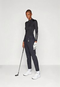 Άτομο στέκεται κρατώντας ένα γκολφ πούτερ, φορώντας μαύρη αθλητική μπλούζα, μαύρο παντελόνι, λευκό γάντι γκολφ, λευκές κάλτσες και λευκά αθλητικά παπούτσια σε μονόχρωμο φόντο.