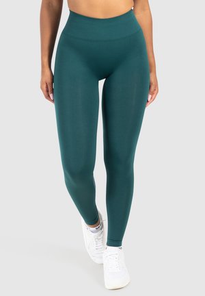 Vrouw in hoogopgetrokken donkergroene leggings en witte sneakers, staand met iets gekruiste benen tegen een lichtgrijze achtergrond.