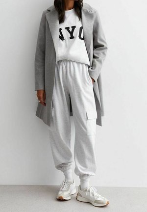 Femme portant un manteau gris par-dessus un sweat-shirt blanc NY, un pantalon de jogging gris clair avec des poches cargo, des chaussettes blanches et des baskets blanches.