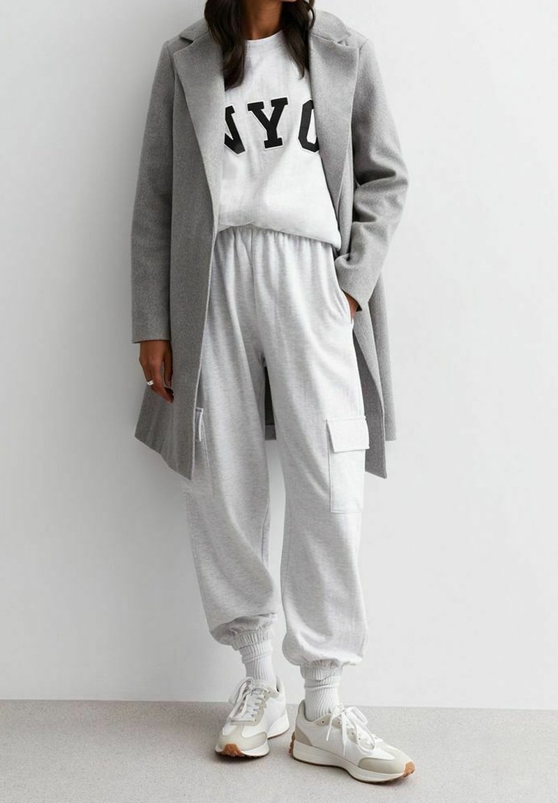 Femme portant un manteau gris par-dessus un sweat-shirt blanc NY, un pantalon de jogging gris clair avec des poches cargo, des chaussettes blanches et des baskets blanches.