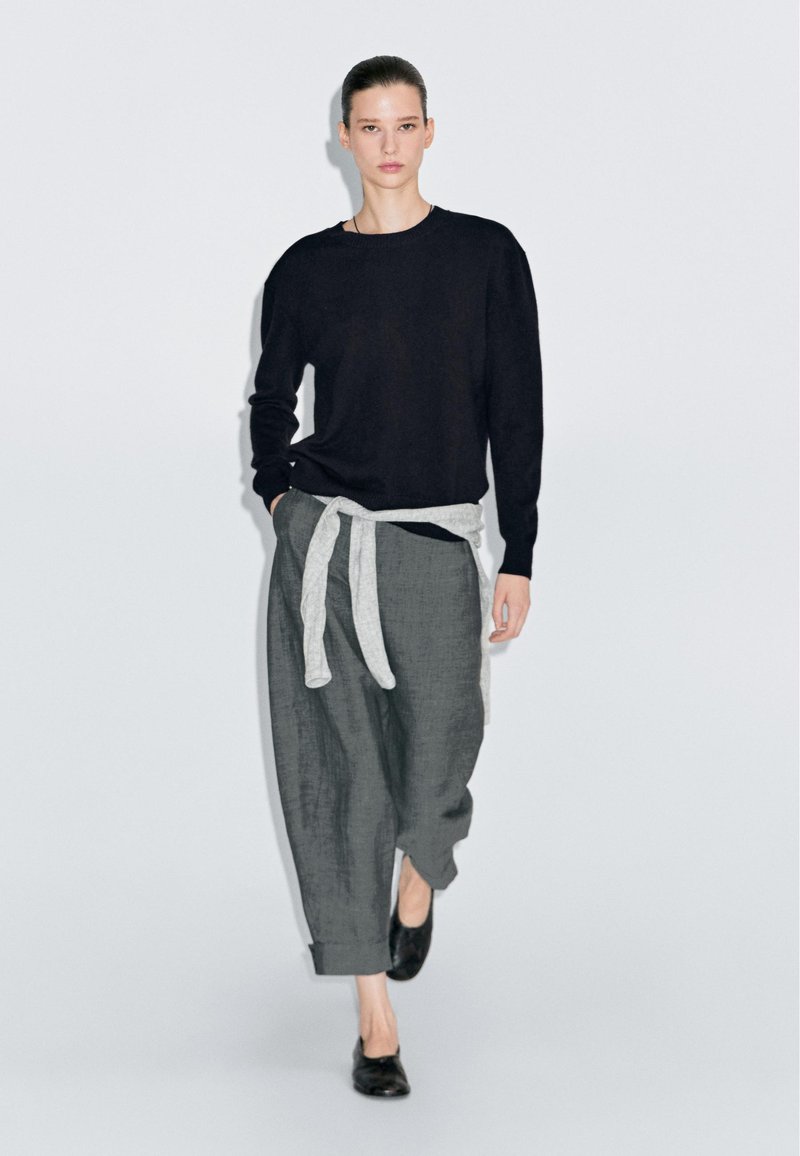 Maglione nero con maniche lunghe, pantaloni grigi legati con una cintura grigia chiara e scarpe nere. Il modello è in piedi contro uno sfondo bianco.