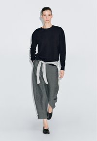 Černý svetr s dlouhými rukávy, šedé kalhoty stáhnuté světle šedým páskem a černé boty. Modelka stojí na bílém pozadí.